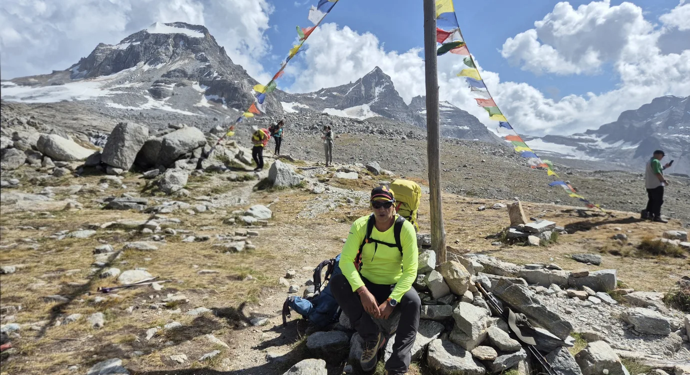 Ο Βαγγέλης κάνει στάση σε αλπικό μονοπάτι στο Gran Paradiso δίπλα σε σημαίες προσευχής, με παγετώνες και κορυφές στο βάθος.