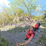 Κατηφορικό κομμάτι της διαδρομής, με την ομάδα να κινείται μέσα στο πράσινο. Πεζοπόροι κατηφορίζουν μονοπάτι μέσα σε δάσος με πλατάνια και βραχώδες τοπίο