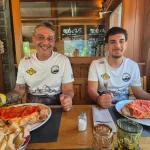 Βαγγέλης και Γιάννης, έτοιμοι για όλα. Ο Βαγγέλης και ο Γιάννης χαμογελούν μπροστά από τις πίτσες τους σε ξύλινο εστιατόριο.