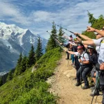 Όλα τα βλέμματα στην κορυφή του Mont Blanc. Η ομάδα στέκεται στο μονοπάτι και δείχνει προς την κορυφή του Mont Blanc, με παγετώδες αλπικό τοπίο να απλώνεται απέναντι.