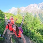 Η Λένα μας, πάντα με χαμόγελο. Η Λένα βγάζει χαμογελαστή selfie με δύο μέλη της ομάδας σε καταπράσινο αλπικό μονοπάτι, με απόκρημνες κορυφές στο βάθος.