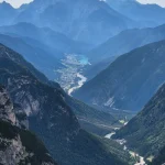 Η κοιλάδα ανοίγεται κάτω από το μονοπάτι πανοραμική θέα σε βαθιά ορεινή κοιλάδα με δασωμένες πλαγιές, οικισμό και γαλαζοπράσινη λίμνη στο βάθος