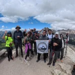 Η ομάδα ψηλά, με χαμόγελα και θέα ομαδική φωτογραφία πεζοπόρων με τη σημαία του BackpackOnGreece σε ψηλό ορεινό σημείο με κορυφές στο βάθος