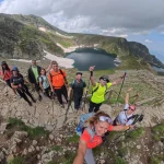 Ανεβαίνουμε μαζί, χαμογελάμε ακόμα περισσότερο η ομάδα ανεβαίνει σε βραχώδες ορεινό μονοπάτι με αλπική λίμνη και χιονούρες στο βάθος