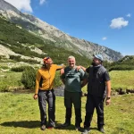 Παρέα, βουνό και χαμόγελα τρία μέλη της ομάδας ποζάρουν χαμογελαστά σε ορεινό λιβάδι με βραχώδεις πλαγιές και ρυάκι στο φόντο