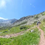 Η ανηφόρα συνεχίζεται, η θέα ανοίγει πεζοπόροι ανεβαίνουν σε ανοιχτό ορεινό μονοπάτι ανάμεσα σε πράσινες πλαγιές, βράχια και ψηλές κορυφές