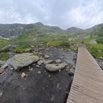 Μια ήσυχη γωνιά μέσα στο βουνό ξύλινο πέρασμα πάνω από ορεινό ρυάκι δίπλα σε αλπική λίμνη, βραχώδεις πλαγιές και πράσινο ορεινό τοπίο