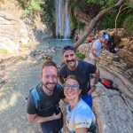 Χαμόγελα με φόντο τον καταρράκτη Τρία μέλη της ομάδας χαμογελούν μπροστά στη φυσική βάθρα και τον καταρράκτη